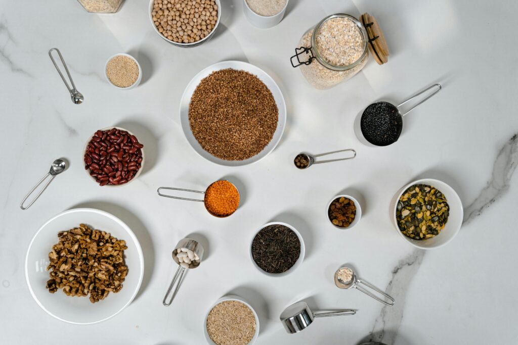 Flat lay of various dried grains, seeds, and beans arranged in bowls and measuring cups on marble.